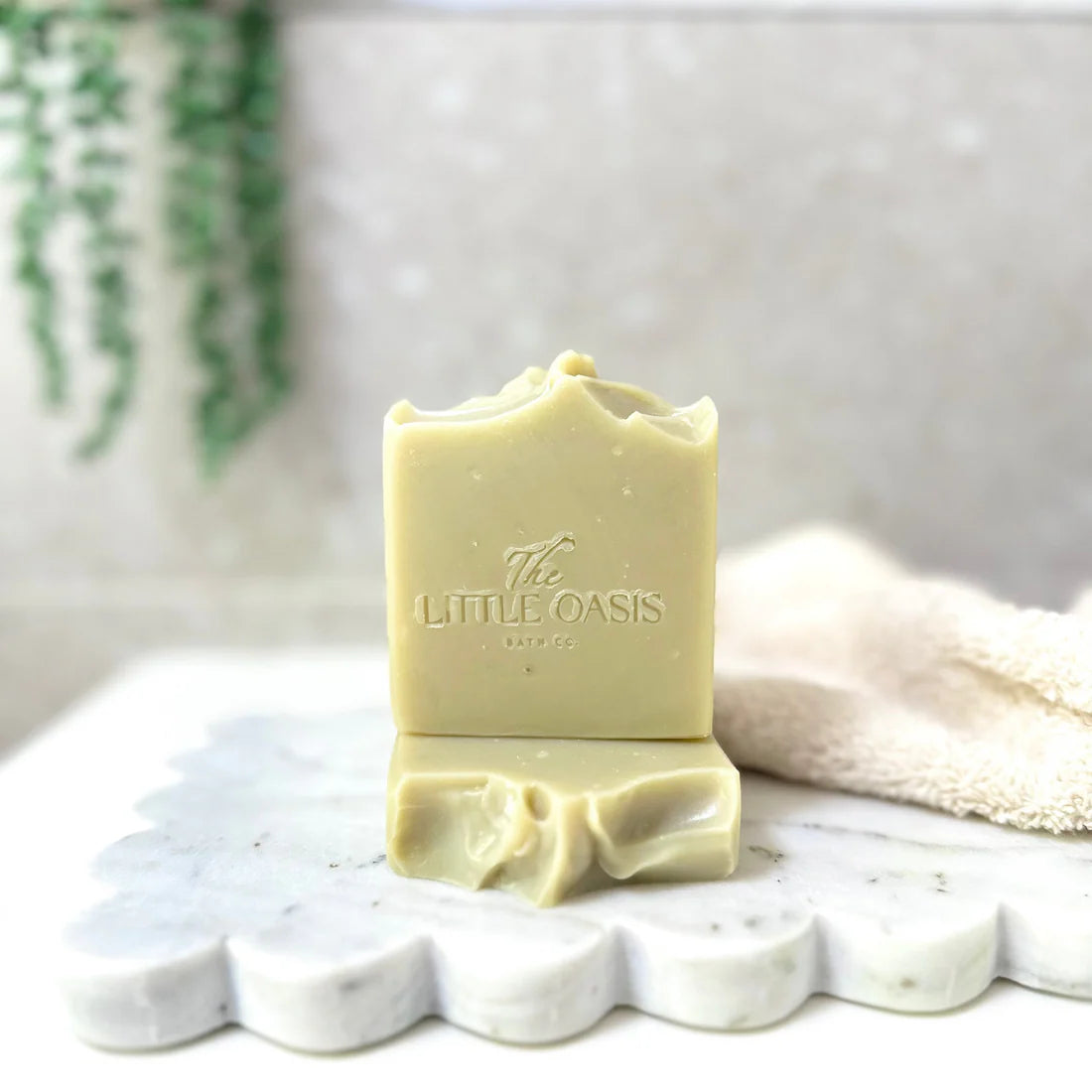 A bar of pale yellow-green handmade soap standing upright on a matching base, with the embossed brand name 'The LITTLE OASIS BARCO' visible on the front. The soap is styled on a scalloped white marble tray with a soft beige towel and a blurred green plant in the background.