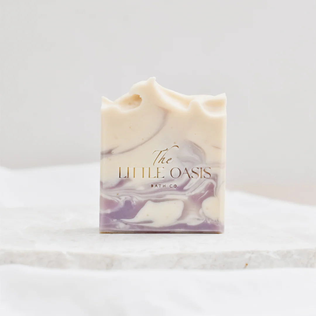 A single bar of handmade soap with cream and lavender-purple marbled swirls, standing upright on a white marble surface. The soap features the brand name 'The LITTLE OASIS BATH CO' printed in elegant gold foil lettering on the front face.