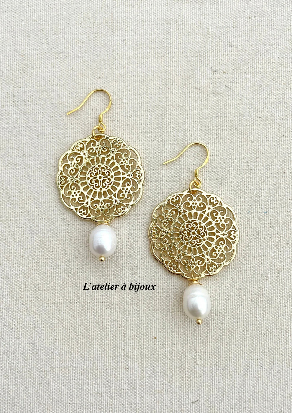 A pair of gold-tone dangle earrings with intricate filigree circular cutouts and a small white pearl drop, displayed on a textured beige fabric background.