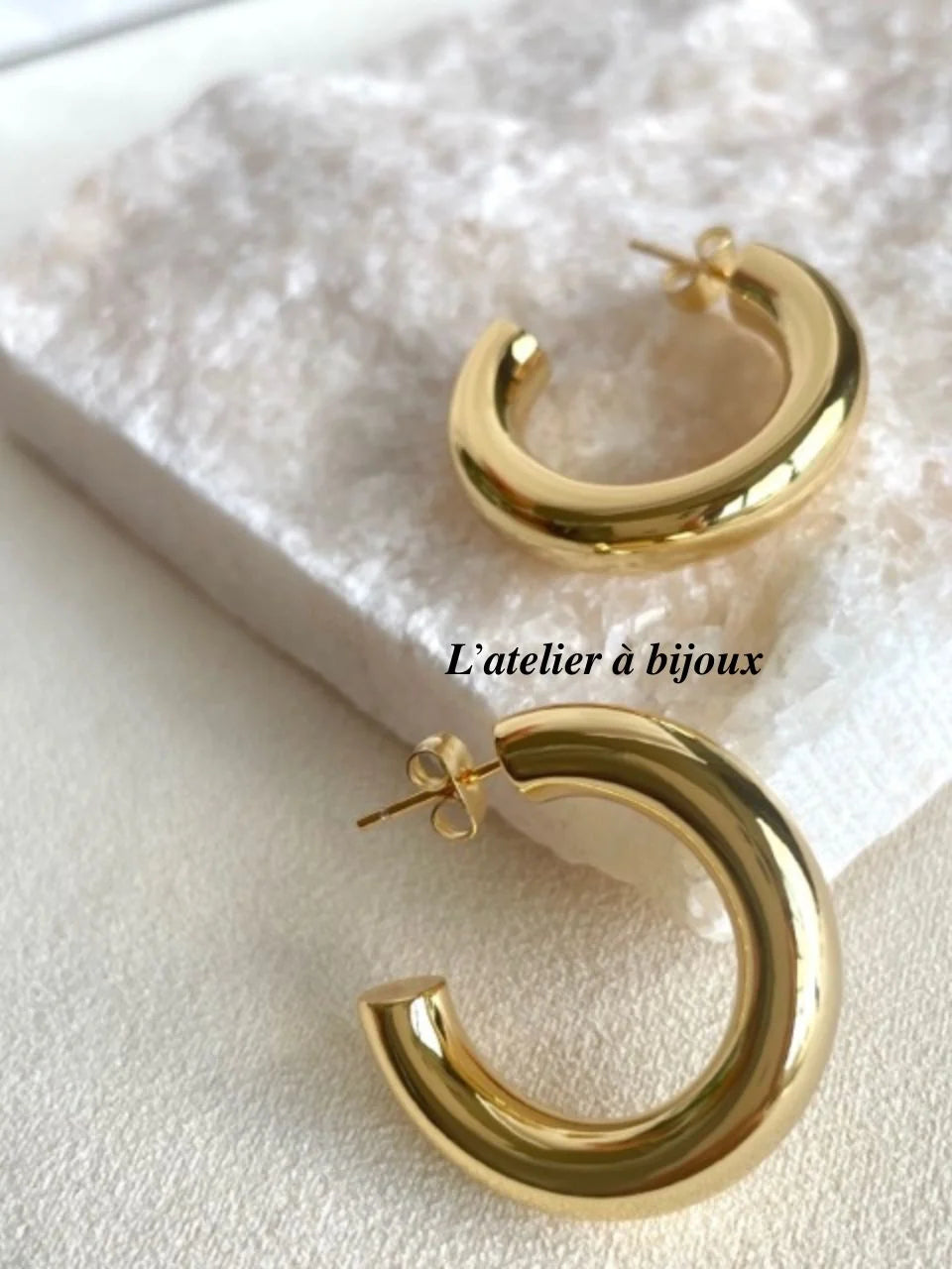 A pair of polished gold-tone hoop earrings displayed on a textured white surface, with a black text watermark reading 'L'atelier à bijoux' across the center.