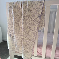 A quilted baby blanket with a lavender floral toile pattern on a cream background, draped over the side of a white wooden crib.