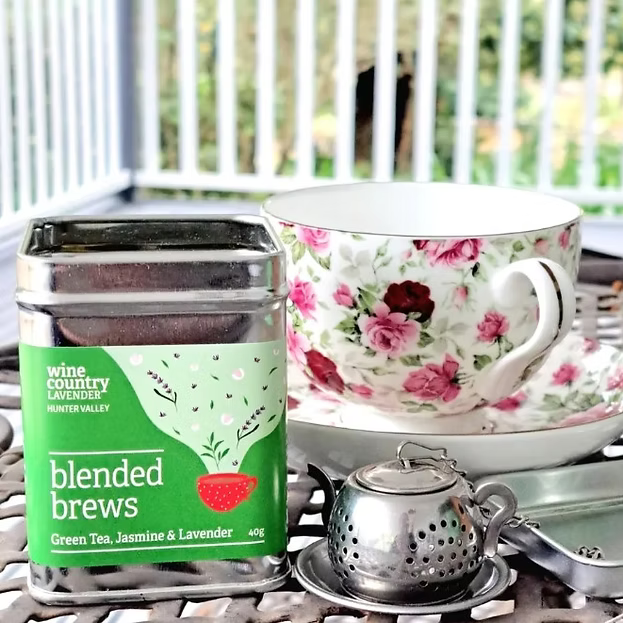 A tin of wine country lavender blended brews green tea sits on a metal patio table next to a floral teacup and saucer with a small metal teapot, with a blurred outdoor patio background.