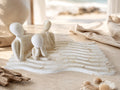 Three abstract white figurines shaped like seated human figures arranged on a wavy rectangular base, styled on a tabletop with linen cloth, shells, and driftwood in a softly blurred beach-like interior setting.