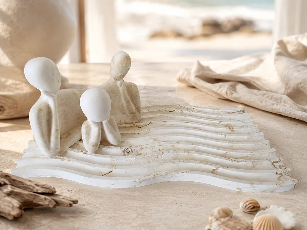 Three abstract white figurines shaped like seated human figures arranged on a wavy rectangular base, styled on a tabletop with linen cloth, shells, and driftwood in a softly blurred beach-like interior setting.
