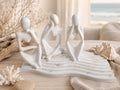 Three white abstract female figurines seated on a matching wave-like base, styled on a tabletop with driftwood, a seashell, coral, and a soft beach-inspired background.