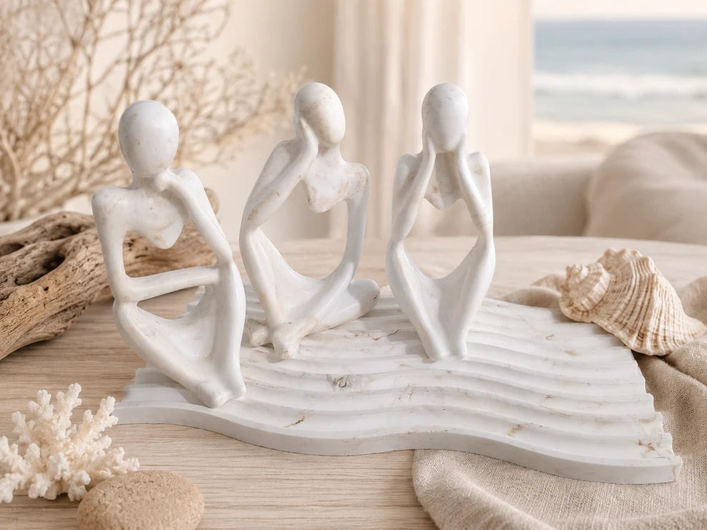 Three white abstract female figurines seated on a matching wave-like base, styled on a tabletop with driftwood, a seashell, coral, and a soft beach-inspired background.