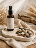 A brown glass spray bottle labeled Sunday Tide Bath Spray stands on a light wood tray with smooth stones, draped over beige fabric with dried flowers and driftwood in the background.