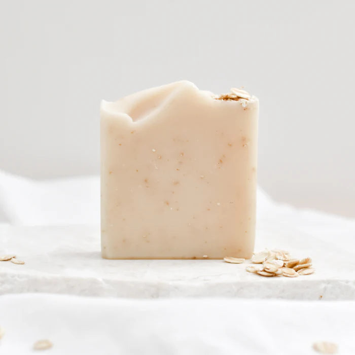 A handmade rectangular bar of oatmeal soap with visible oat flakes and tiny specks, standing on a white marble slab with scattered oats around it.