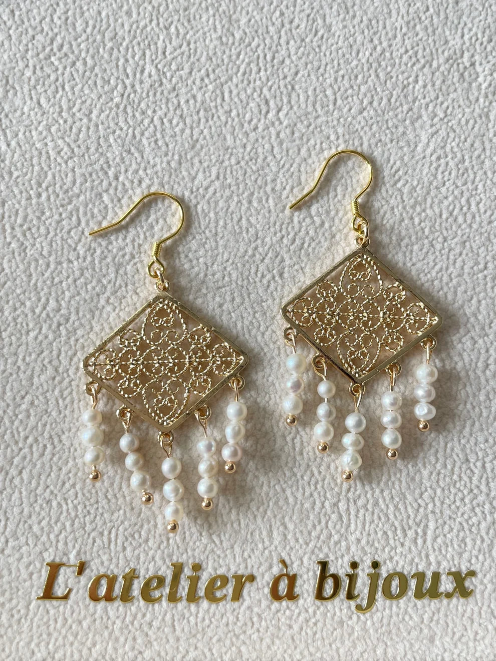A pair of gold-tone dangle earrings with ornate filigree square drops and dangling pearl beads, displayed on a textured light background with the text 'L'atelier à bijoux' at the bottom.
